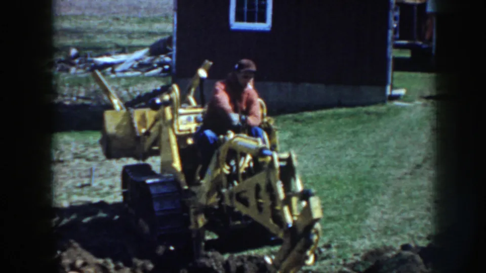 In 1958 Nebraska footage, a man in a red jacket and cap sits on a yellow tracked excavator beside a dark farm outbuilding.