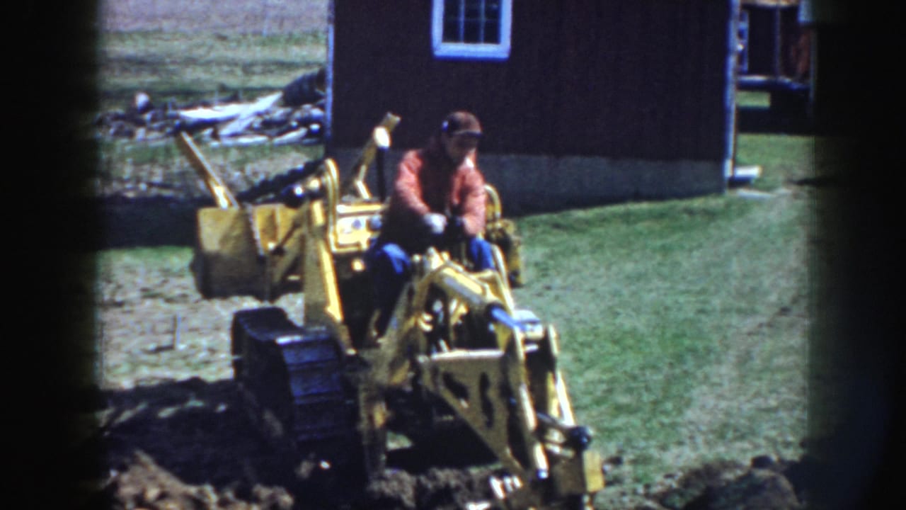 The seated operator works the yellow excavator controls near a dark outbuilding and grassy yard in the Nebraska home-movie scene.