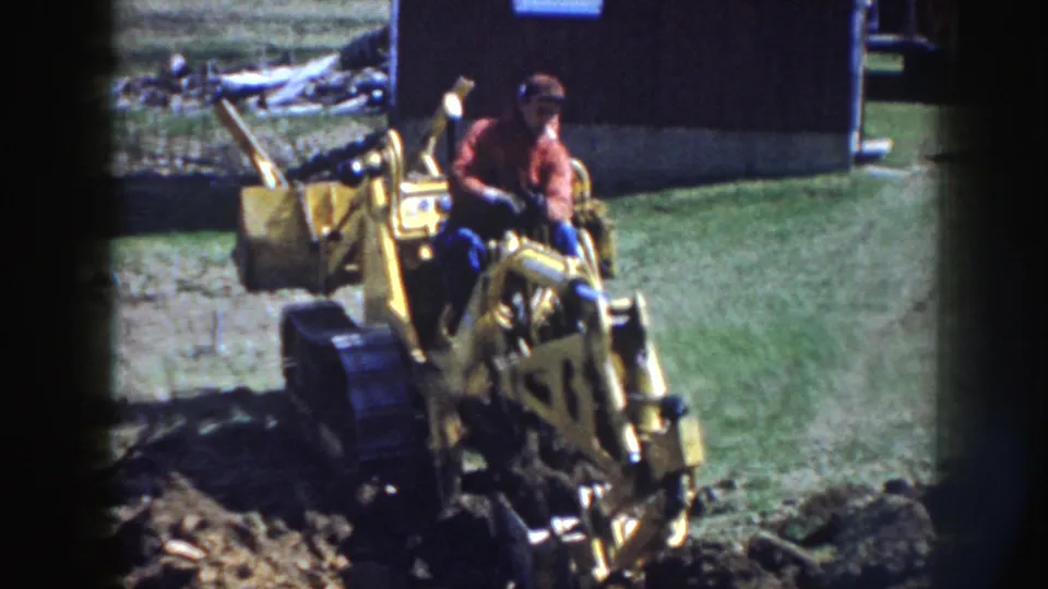 A man in work clothes continues operating the yellow tracked excavator, with dug soil in the foreground and a dark outbuilding behind him.
