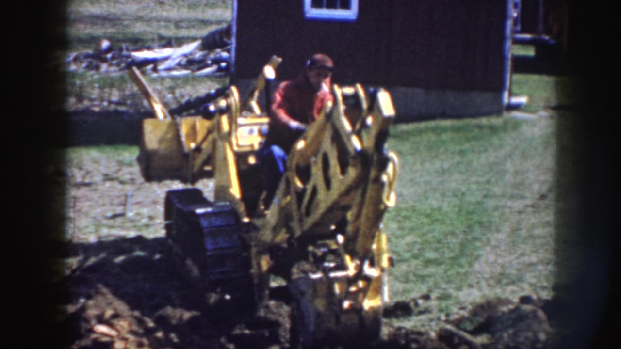 In rural Nebraska 1958 footage, a man operates a yellow backhoe in a grassy yard beside a dark wooden outbuilding.