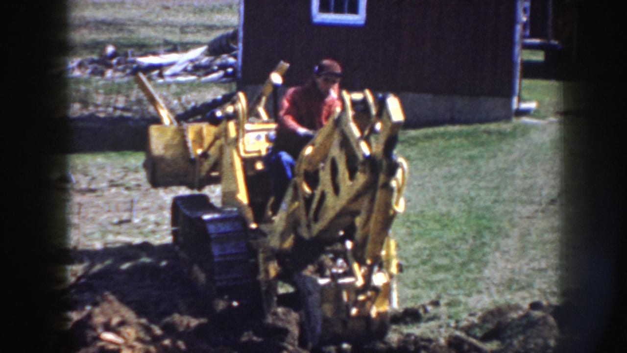 The operator sits at the controls of a yellow backhoe while digging soil in a grassy rural yard.
