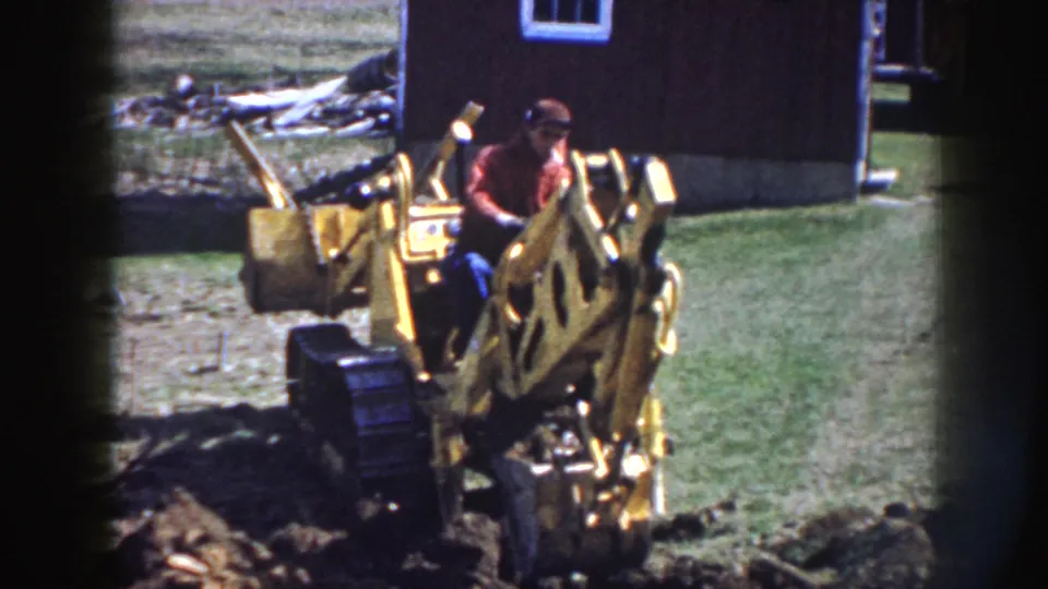 The operator sits at the controls of a yellow backhoe while digging soil in a grassy rural yard.