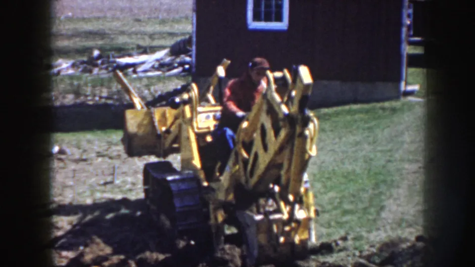 A man in work clothes operates the yellow backhoe close to a trench, with a dark shed and open yard behind him.