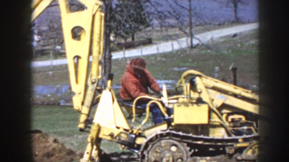 Side view shows the backhoe boom and seated operator working near a dirt pile, with yard and road visible beyond.