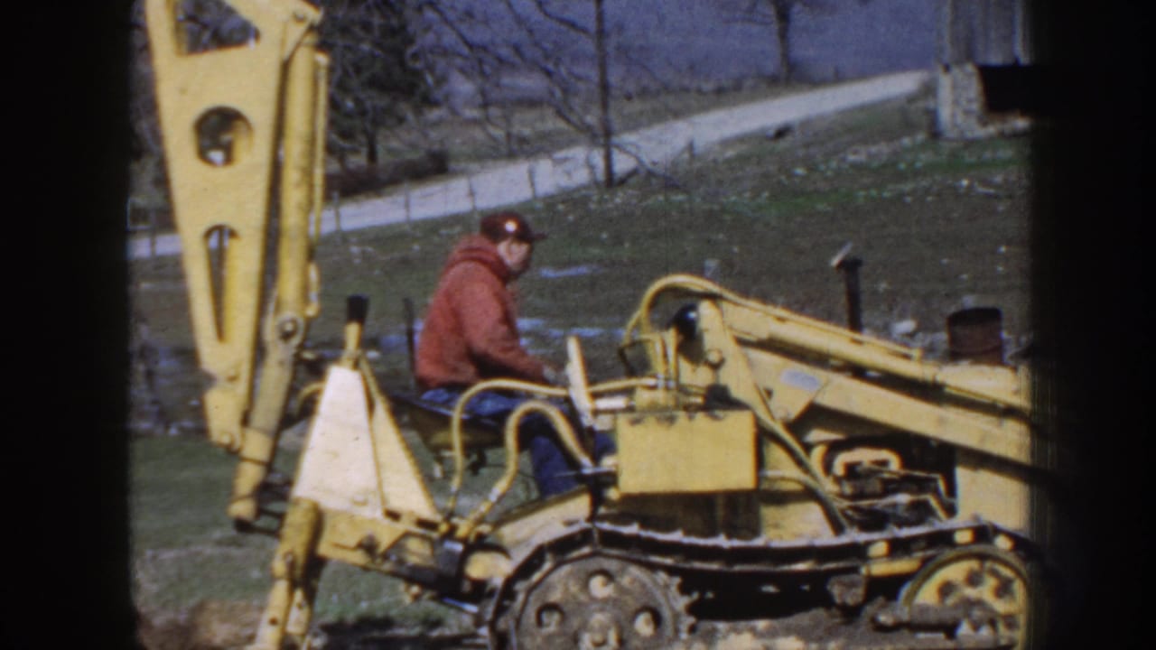 A man in work clothes sits at the controls of a yellow tracked backhoe on rural Nebraska property in 1958.