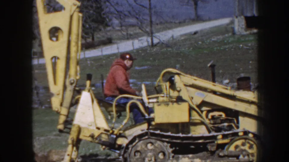 A man in work clothes sits at the controls of a yellow tracked backhoe on rural Nebraska property in 1958.