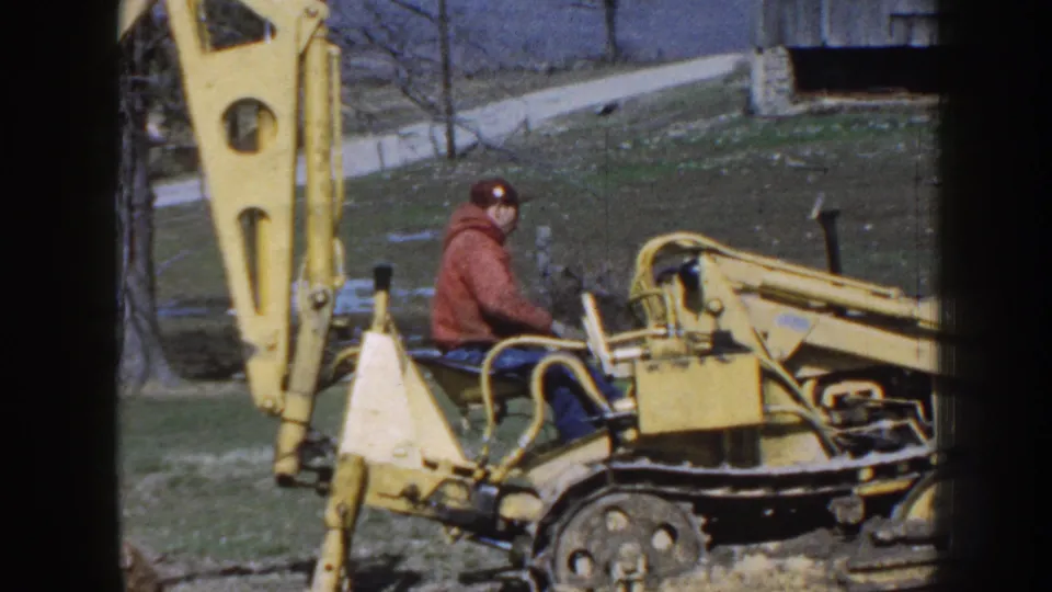 Side view of a yellow tracked backhoe and seated operator, with bare trees and rural ground visible behind them.