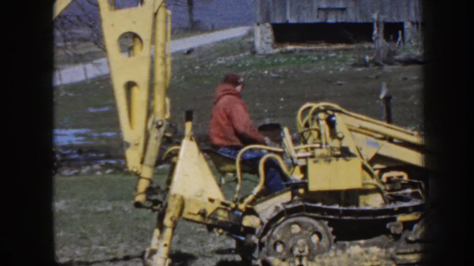 A man on a yellow backhoe works in a grassy Nebraska field, with a dark barn-like structure and road behind him.