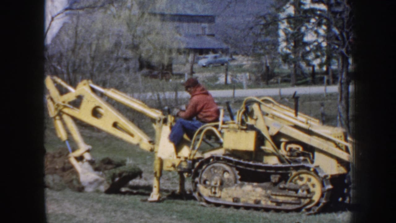 1958 Nebraska home-movie frame showing a man in a cap using a yellow tracked backhoe beside churned soil, with a house and trees beyond.