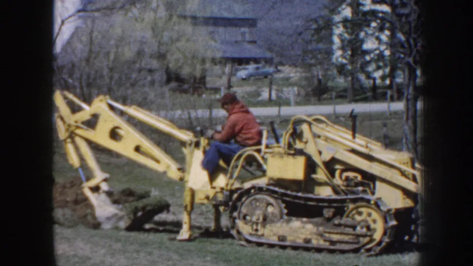 1958 Nebraska home-movie frame showing a man in a cap using a yellow tracked backhoe beside churned soil, with a house and trees beyond.