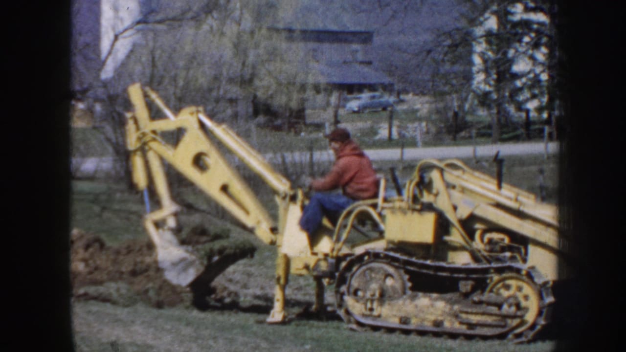 The operator sits on the yellow tracked backhoe as the bucket lifts soil on a Nebraska rural property, with a road and house in the background.