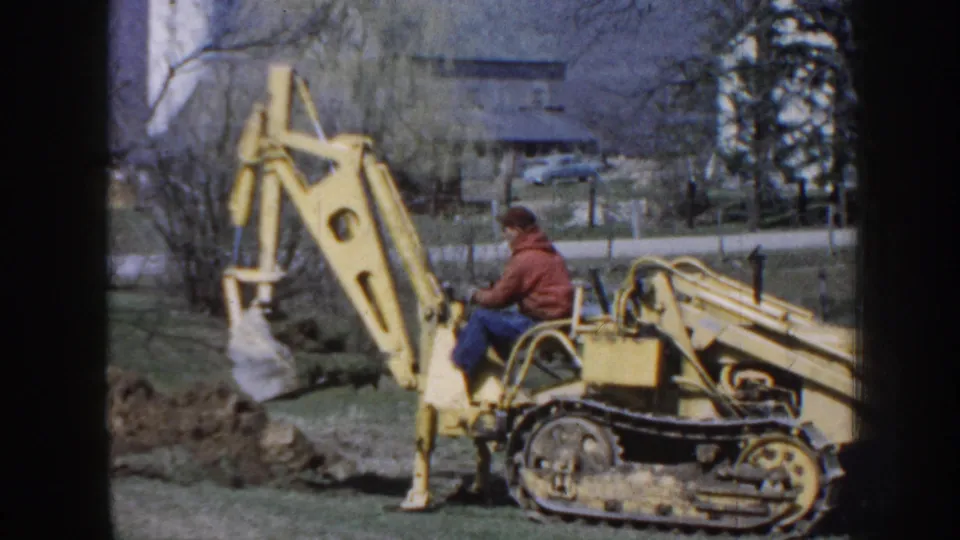 A 1958 Nebraska 8mm frame shows the backhoe arm raised high while the man in a red jacket works near disturbed soil and trees.
