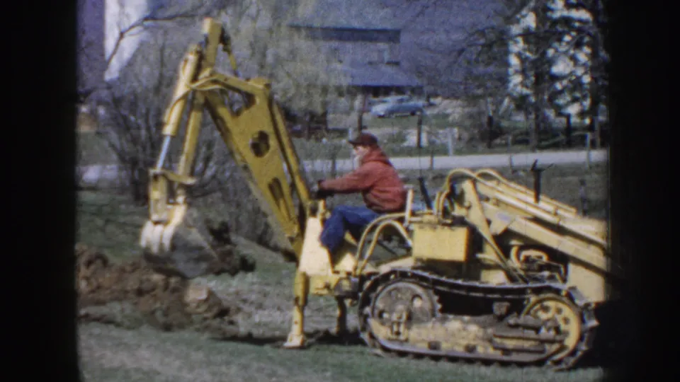 The man guides the yellow tracked backhoe with its bucket suspended over a dug area, filmed in soft 8mm color on a Nebraska property.
