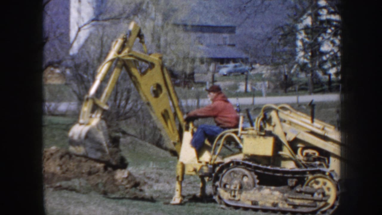 1958 Nebraska home-movie frame showing a man operating a yellow tracked excavator near a dirt pile, with farm buildings and trees behind him.