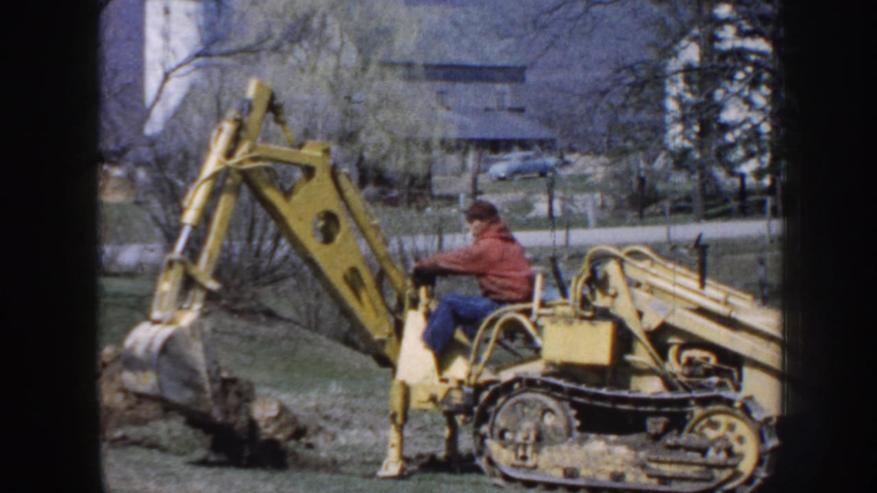 Yellow excavator with seated operator works beside a mound of earth near farm buildings.