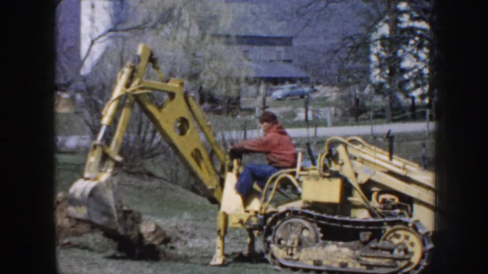 1958 Nebraska frame of a seated operator using a yellow excavator on open grass, with a farmhouse, trees, fence, and silo in the background.
