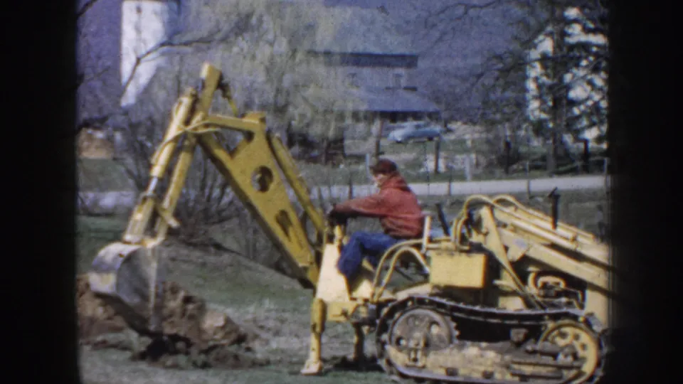 1958 Nebraska home-movie frame showing the excavator bucket near the ground as the operator digs on a rural property.