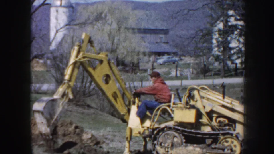 1958 Nebraska frame of a yellow tracked excavator beside a dug area, with the operator seated and rural buildings behind the work site.
