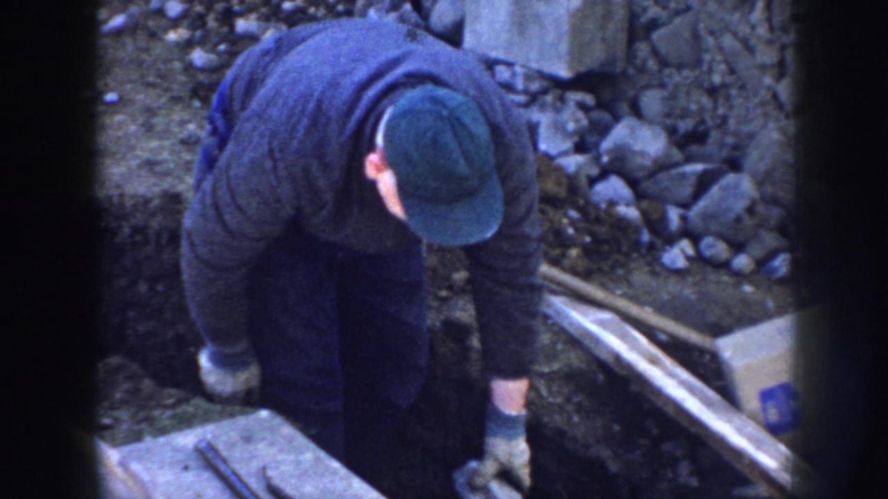 In 1958 Nebraska home-movie footage, a gloved man in a green cap bends over beside a trench at a small construction site.