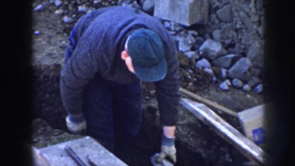 In 1958 Nebraska home-movie footage, a gloved man in a green cap bends over beside a trench at a small construction site.