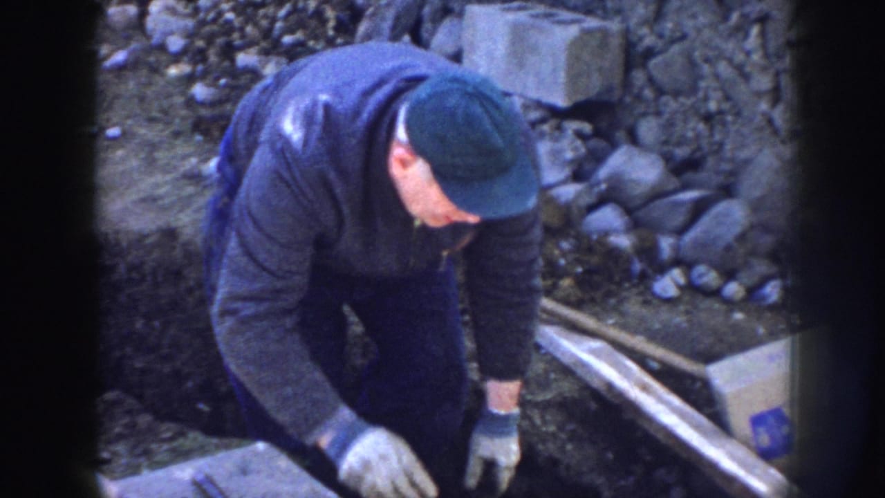 Worker reaches down beside a trench, with rocks, boards, blocks, and a marked box in view.