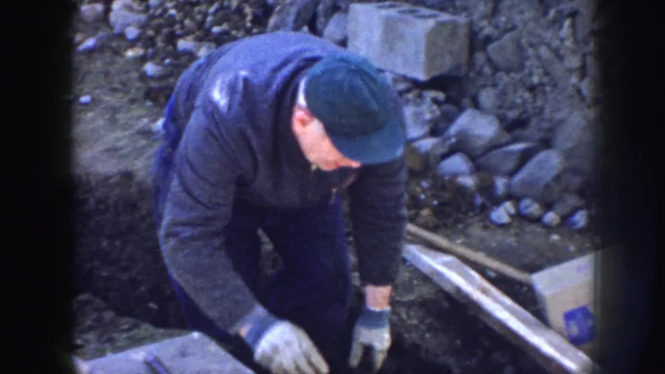A gloved worker at a Nebraska construction site reaches toward materials beside a trench, with a marked cardboard box visible nearby.