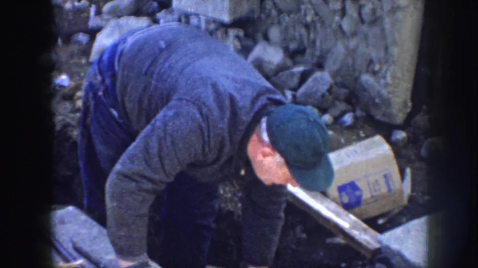 The camera looks down as the worker bends over the construction area, surrounded by stones, lumber, concrete, and a marked box.