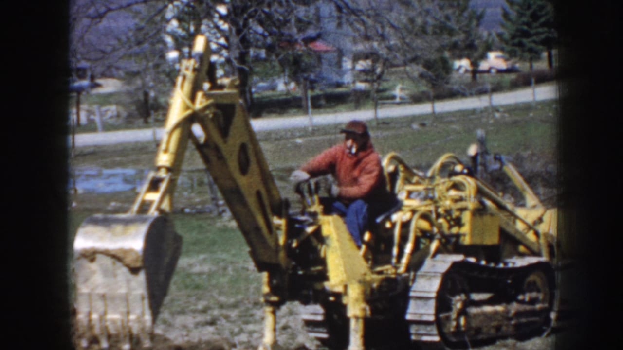 Man in red jacket operating a yellow tracked excavator in a grassy yard.