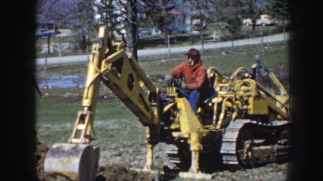 Yellow excavator with seated operator viewed through a dark window-like frame.