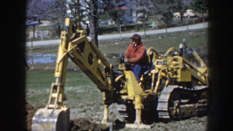 Nebraska 1958 home-movie still of a man at the controls of a yellow tracked excavator with the bucket low to the ground.