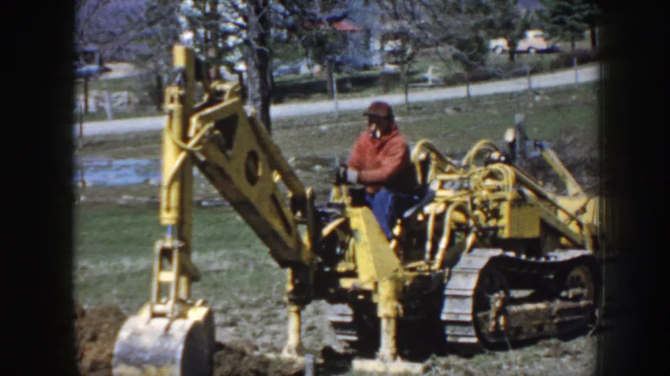Nebraska 1958 frame with a side view of the yellow excavator, seated operator, grassy yard, road, and parked or passing car beyond.