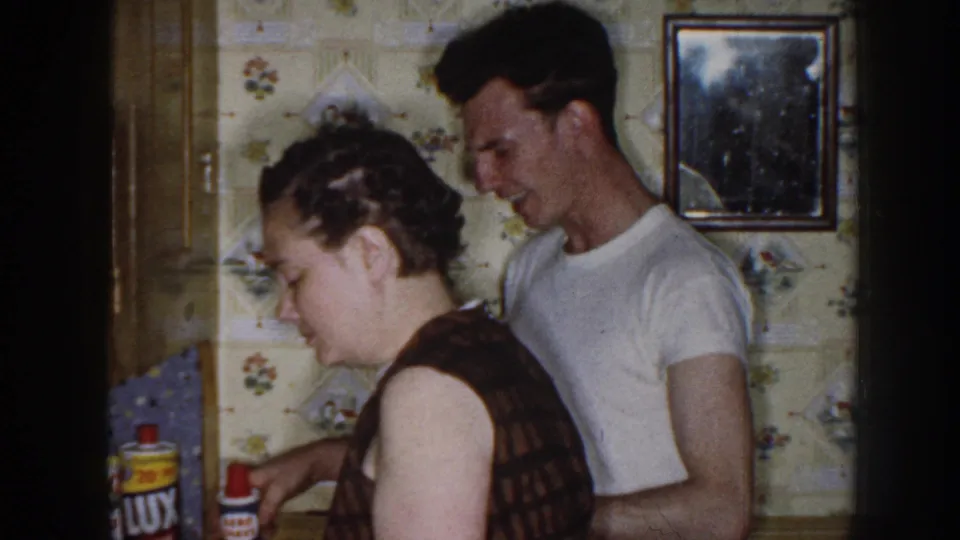 A woman in a dark patterned dress and a man in a white t-shirt stand close in a private Nebraska kitchen, with labeled cleaning products nearby.