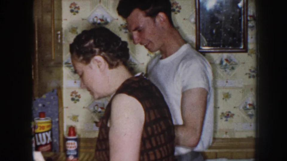 Two adults stand close together in a 1958 Nebraska kitchen interior, with patterned wall covering, a framed picture, and household product labels visible.