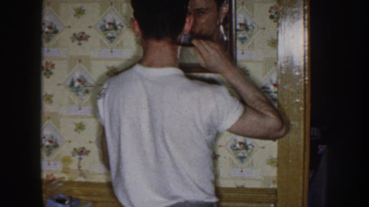 In 1958 Nebraska home-movie footage, a man in a white T-shirt combs his hair before a mirror inside a room with floral wallpaper.