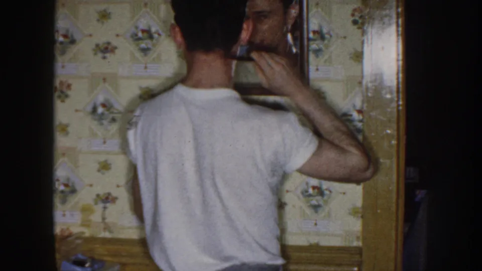 In 1958 Nebraska home-movie footage, a man in a white T-shirt combs his hair before a mirror inside a room with floral wallpaper.