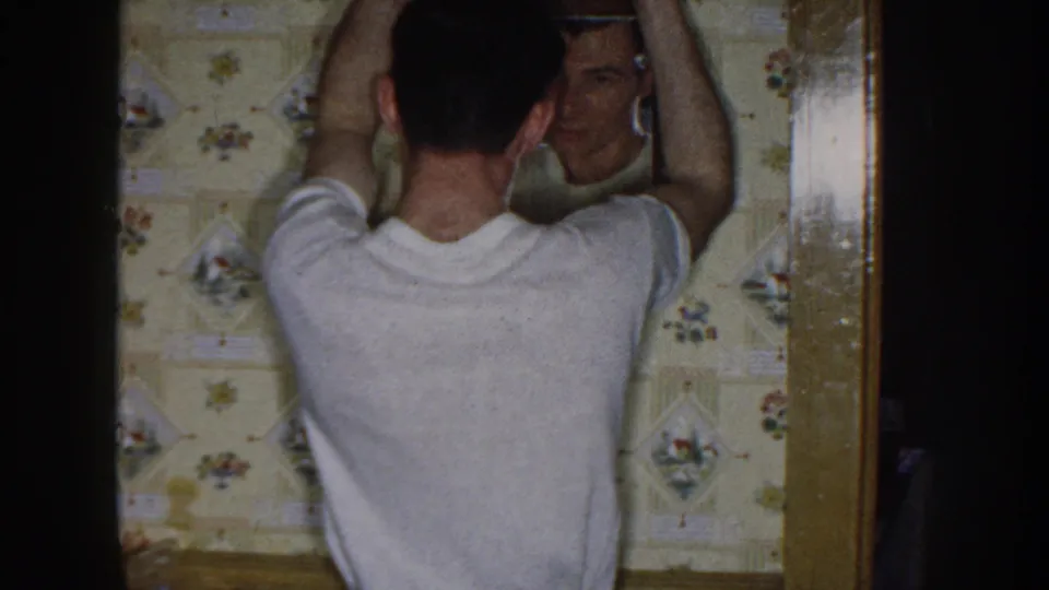 The indoor sequence continues as the man works on his hair before a mirror in a floral-wallpapered private room.