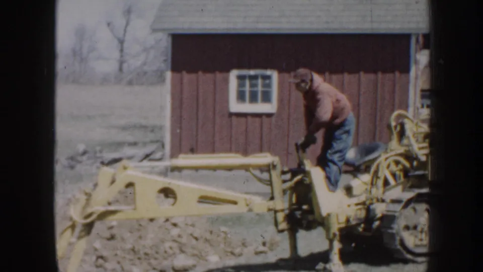 Outdoors in rural Nebraska, an adult appears to work on yellow farm equipment near a red barn and bare trees.
