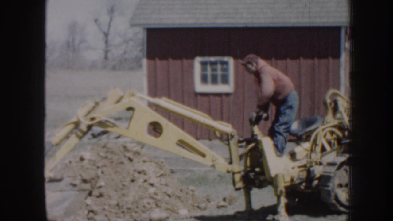 Backhoe arm extends across a dirt pile while the operator stands near the controls.