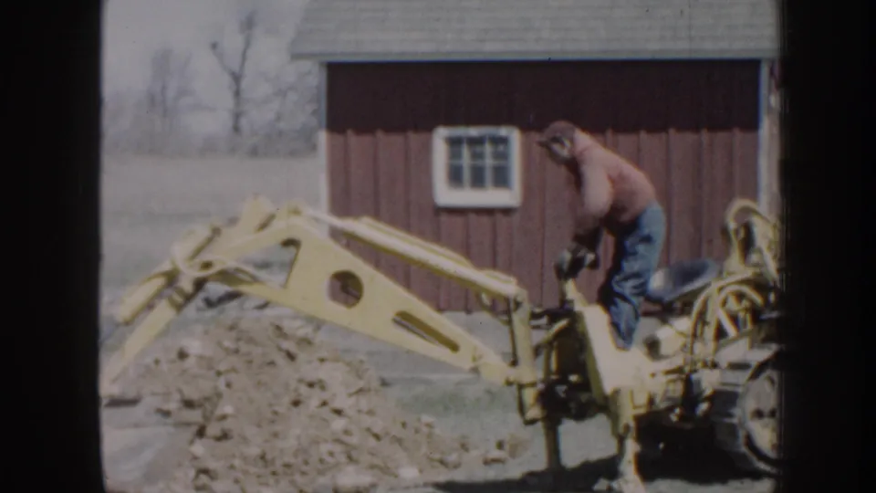 The yellow backhoe arm reaches over excavated dirt as the adult operator works beside a red rural outbuilding in Nebraska.