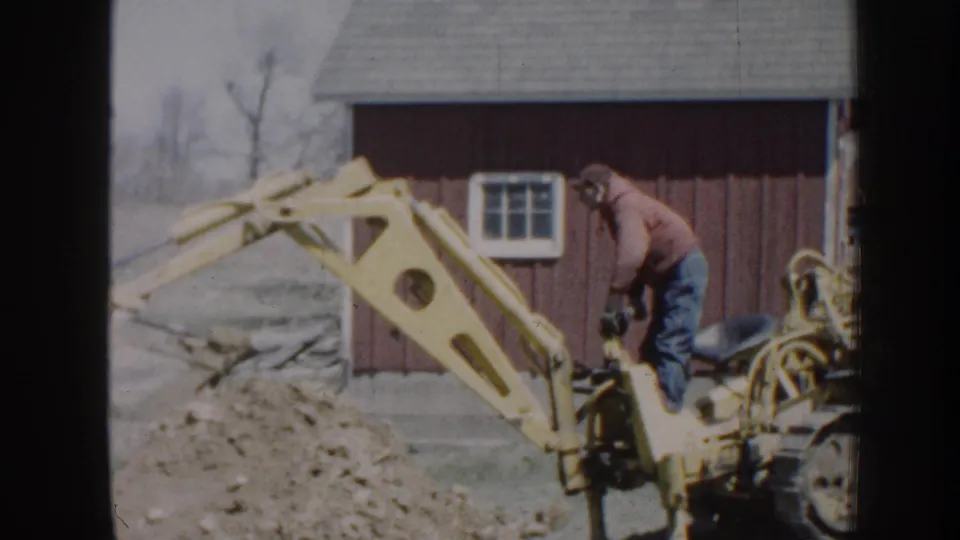 A 1958 8mm frame shows the backhoe boom raised beside a red outbuilding as the operator continues digging work.