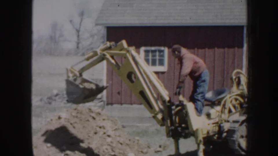 The backhoe bucket is lifted near the end of the short 1958 Nebraska scene, with the operator and red outbuilding still visible.