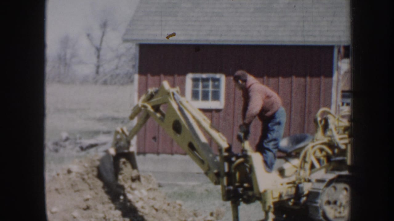 Backhoe bucket lowered into dirt while a man stands at the controls near a red barn.