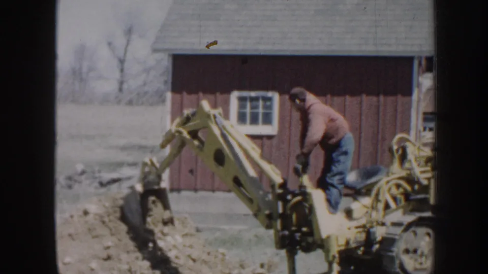 1958 Nebraska farm footage frame with the backhoe bucket cutting into soil as the operator stands on the machine.