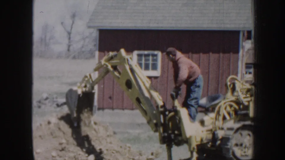 1958 Nebraska 8mm frame showing a hooded man operating a yellow backhoe with the bucket lifting dirt beside a red barn.