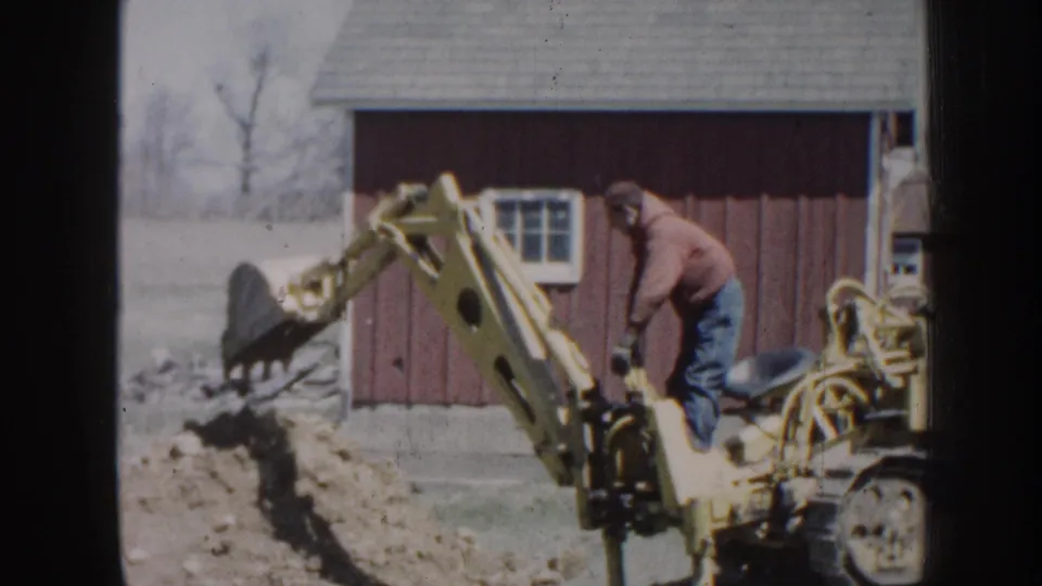1958 Nebraska farm frame with a small yellow backhoe raising its bucket as a man in a red hooded sweatshirt operates it.