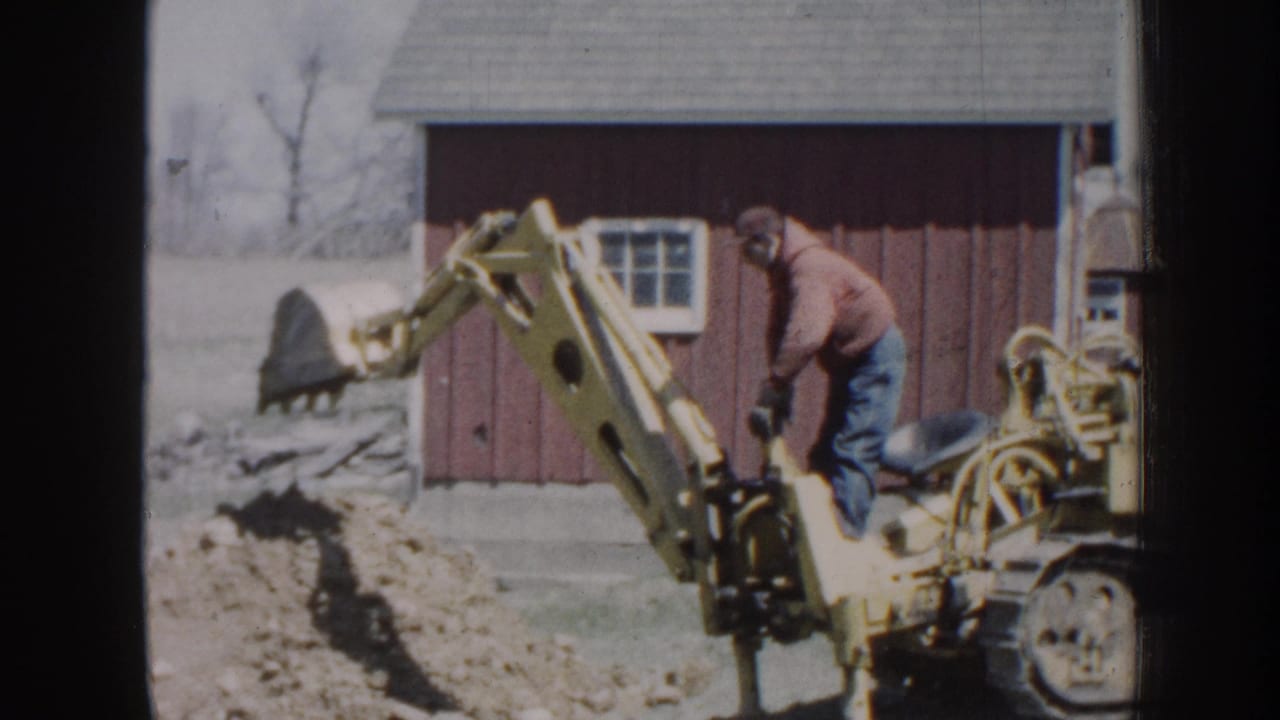 1958 Nebraska home-movie frame showing a man in a jacket operating a yellow excavator near a red outbuilding and disturbed earth.