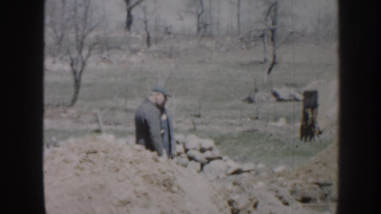 Man in a dark jacket standing near rocks and dug soil in an open field.