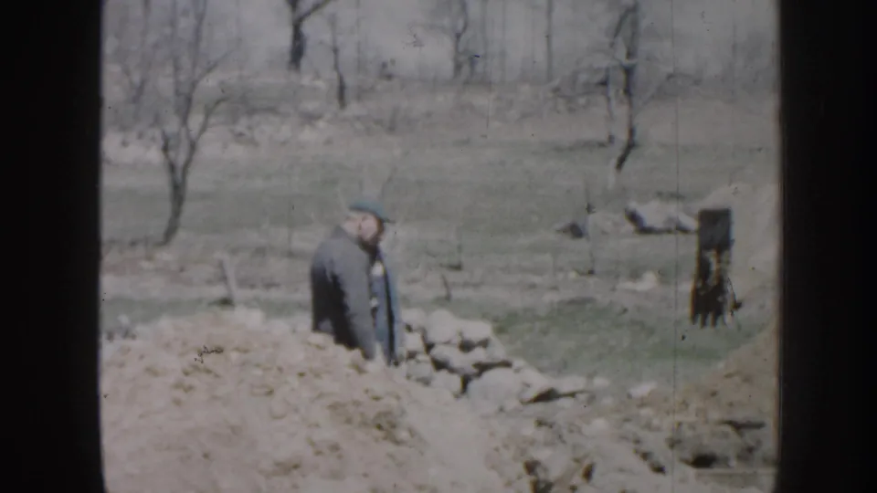 1958 Nebraska rural construction frame with an adult man standing near rocks, excavated earth, grass, and leafless trees.