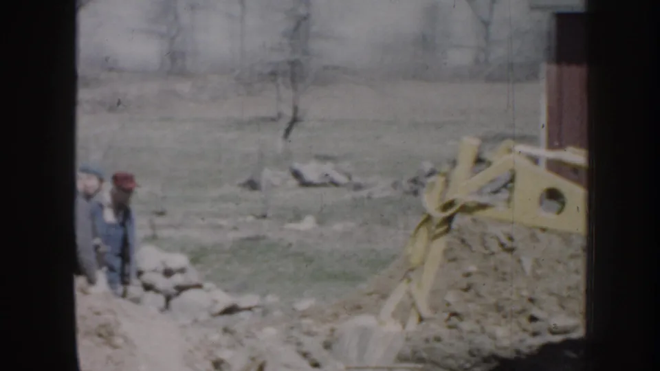 1958 Nebraska home-movie frame showing two men by a trench with a yellow excavator arm, rocks, grass, and bare trees nearby.