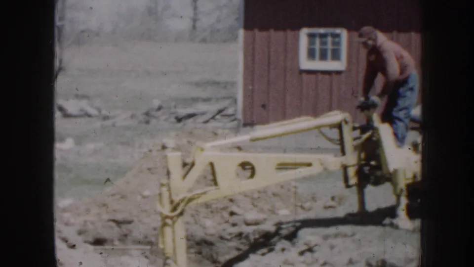 1958 Nebraska frame with a yellow excavator arm, the operator at right, churned soil, and a red outbuilding with a white window.